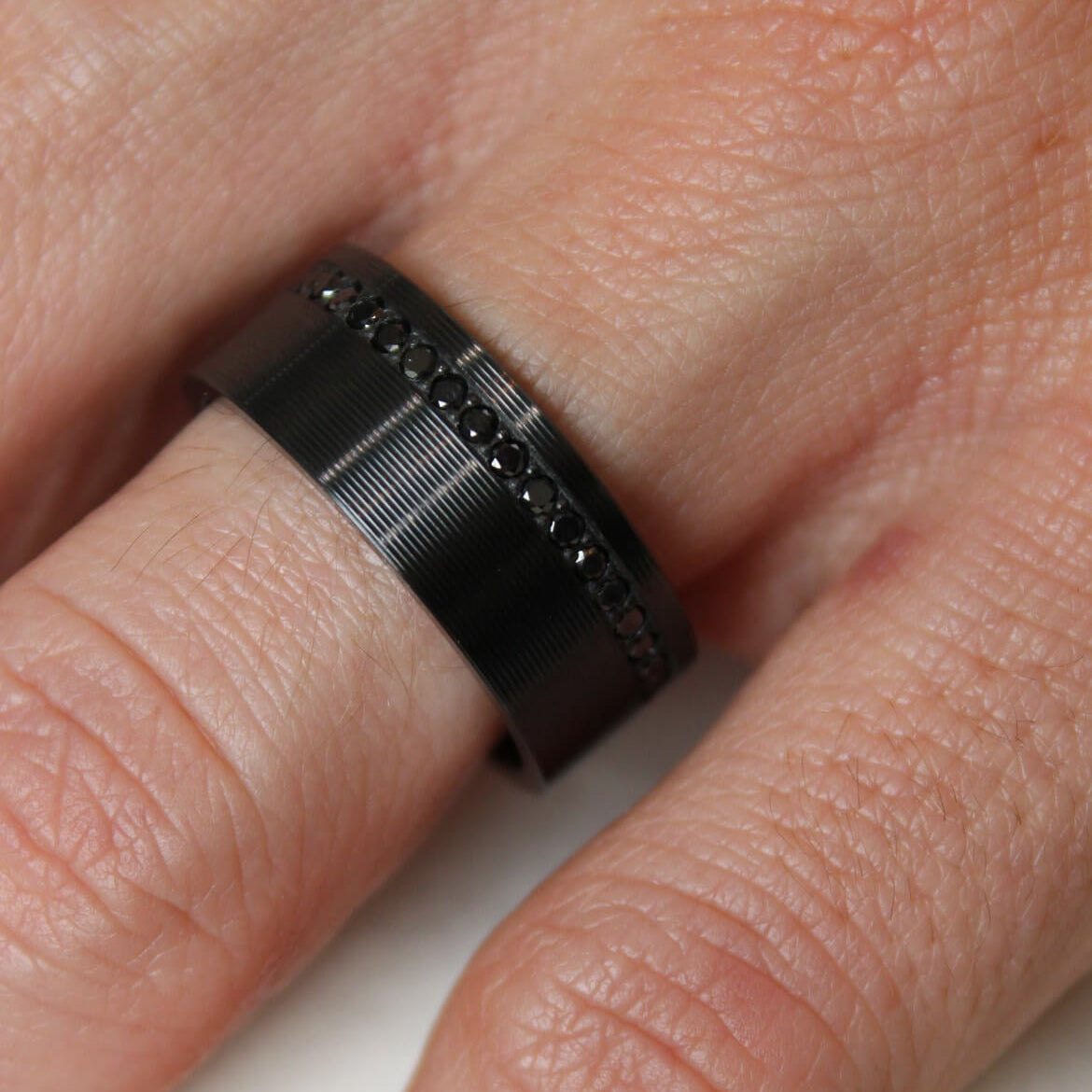 Close-up of a hand wearing a black zirconium men's wedding ring with black lab grown diamonds and a ribbed finish on a white background