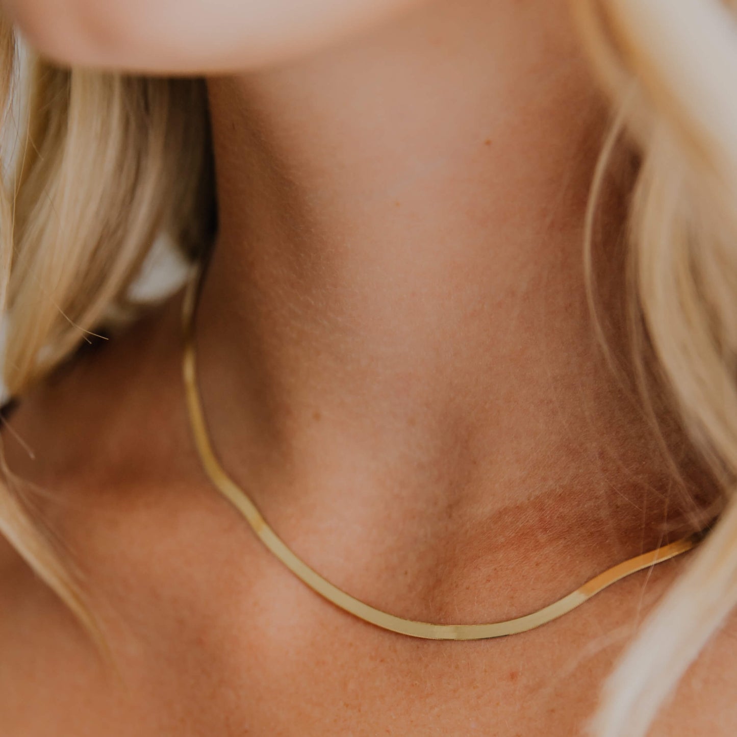 Close-up of a person wearing a thin gold herringbone necklace with blurred background