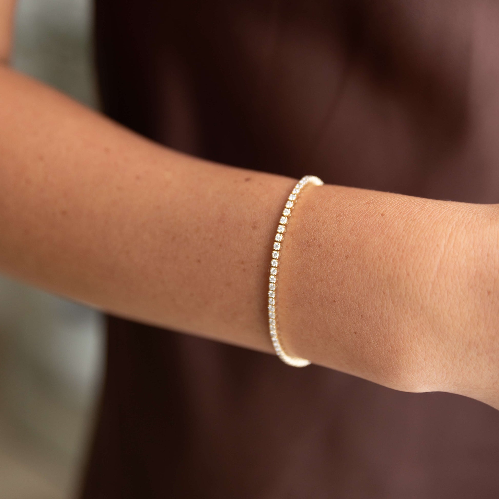 Close-up of a woman's arm wearing a 14K yellow gold diamond tennis bracelet on a blurred background