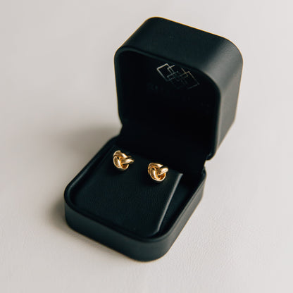 14K Gold love knot earrings in a black SHANNON jewelry box on a light gray background
