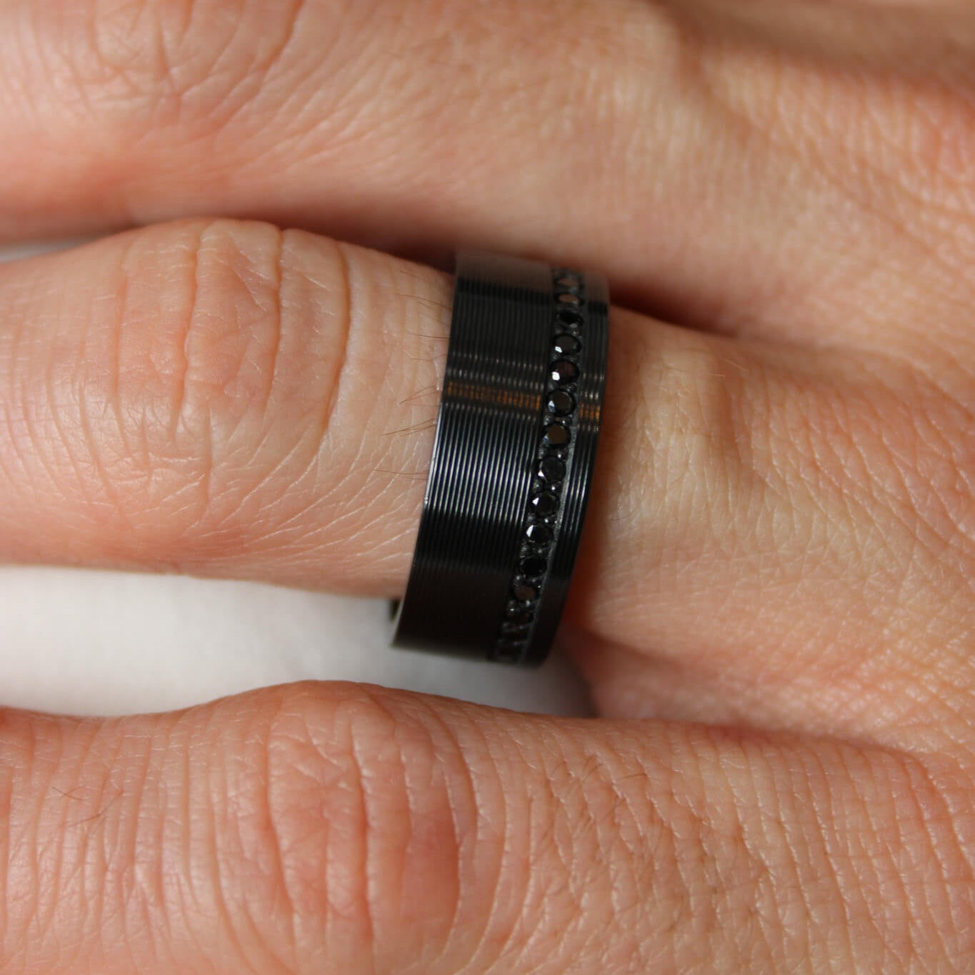 Close-up of a hand wearing a black zirconium men's wedding ring with black lab grown diamonds and a ribbed finish on a white background